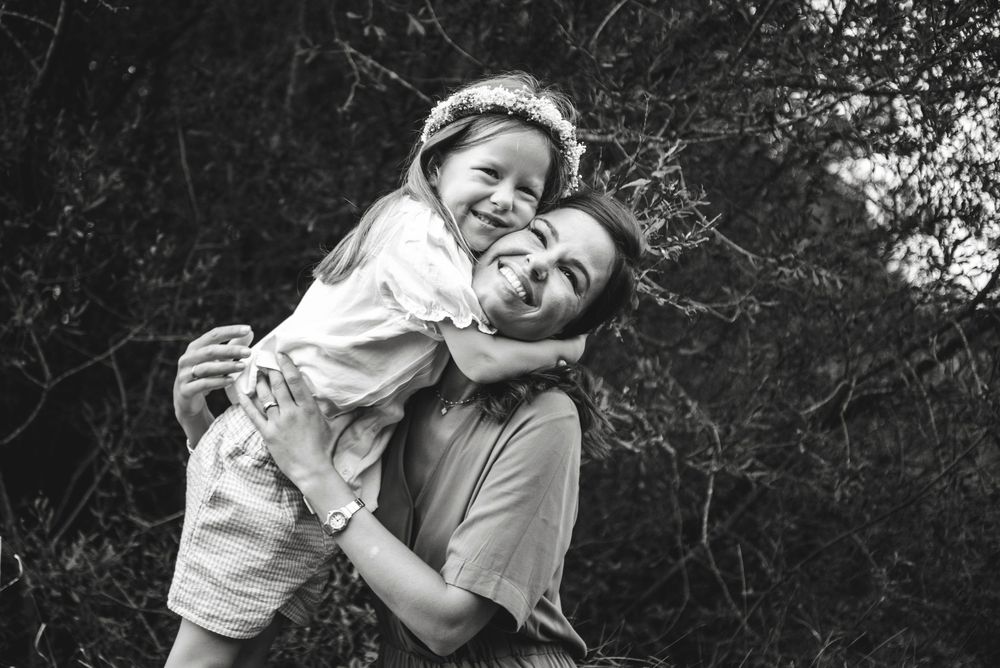 Black and white photo of a smiling mother carrying her happy young daughter outdoors among trees.
