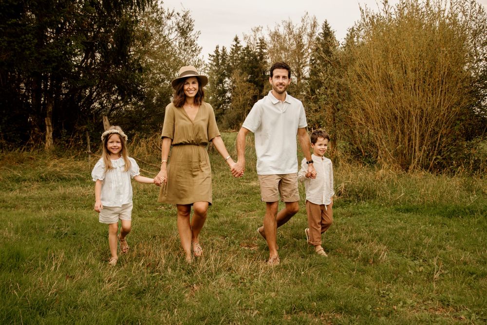Family of four walking barefoot in a green meadow, parents holding hands with two young children on a sunny day.