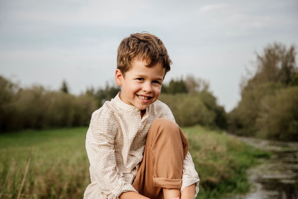 Smiling young boy sitting outdoors near a stream, wearing a white shirt and tan pants, surrounded by green nature.