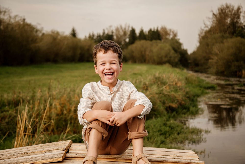Joyful young boy sitting cross-legged on wooden dock, laughing outdoors near a pond with lush green meadow.