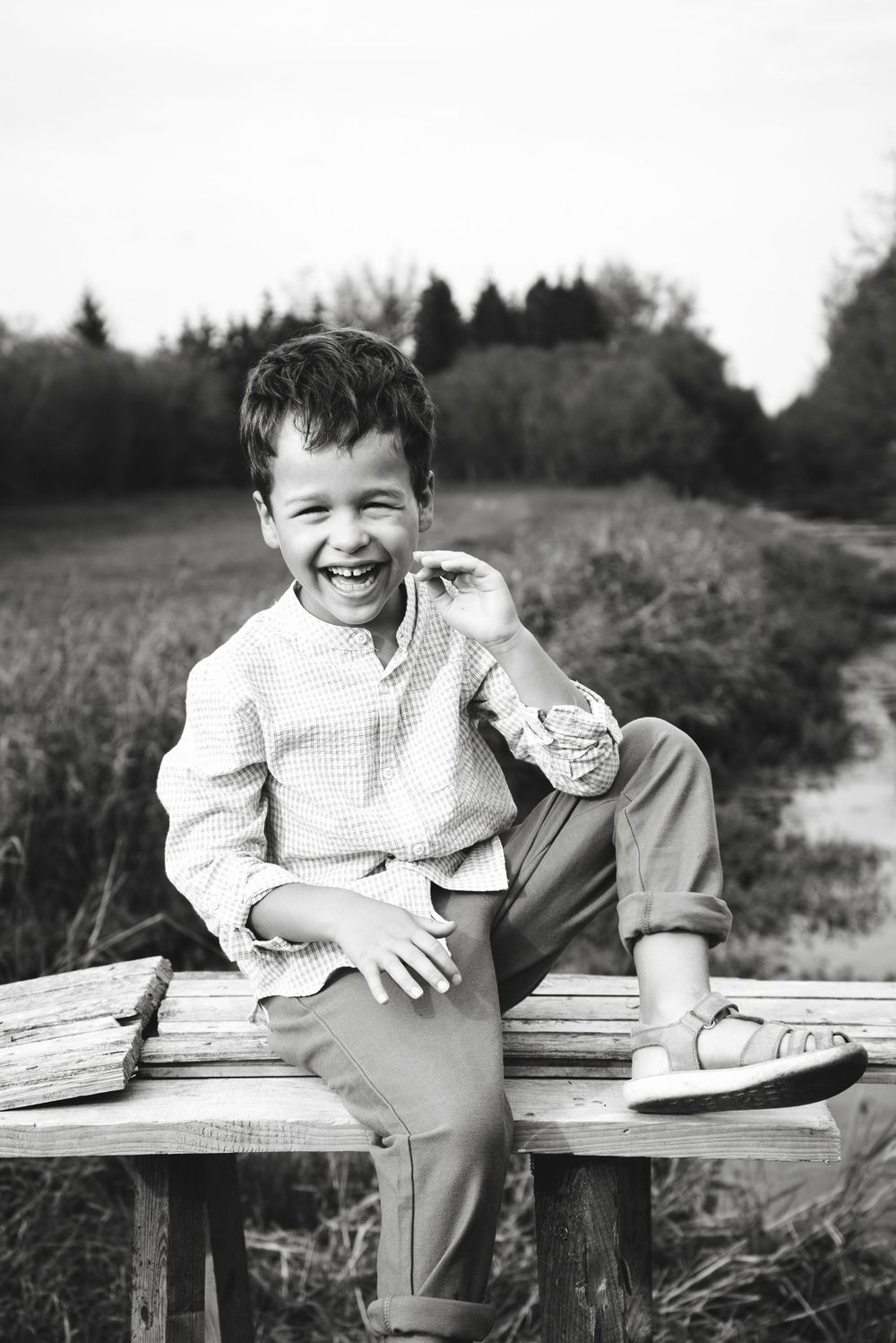 Black and white photo of a laughing boy sitting on a wooden bench in a rural field setting.