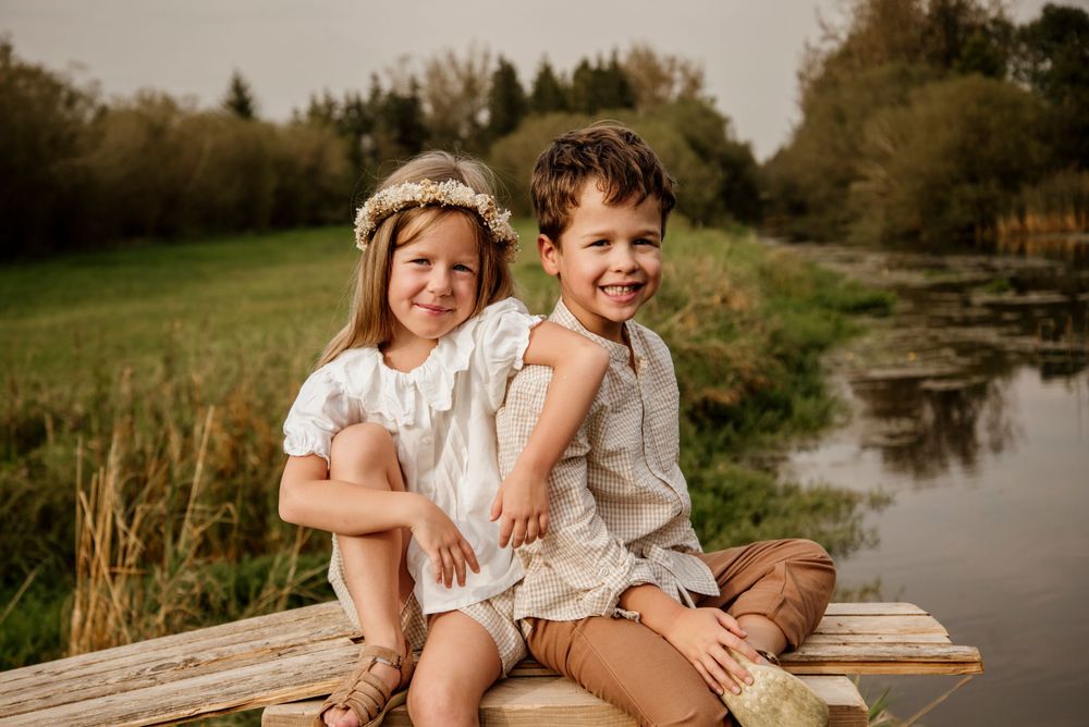 Two young children sitting on a wooden dock by a pond, smiling, girl wearing floral crown, boy in linen outfit.