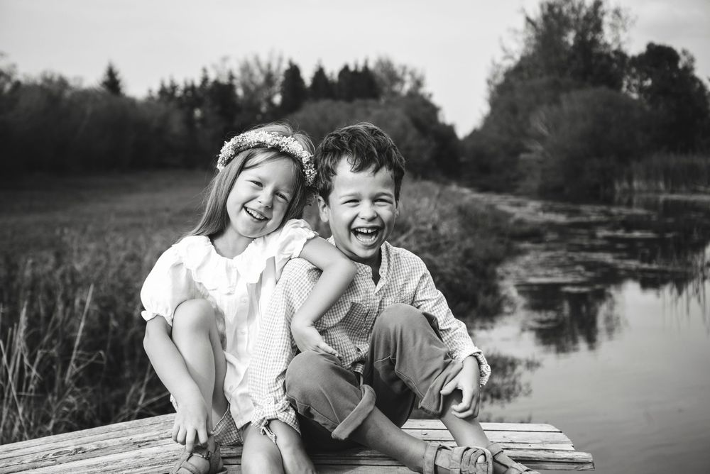 Two laughing children sit together on a wooden dock by a pond in a black and white outdoor portrait.