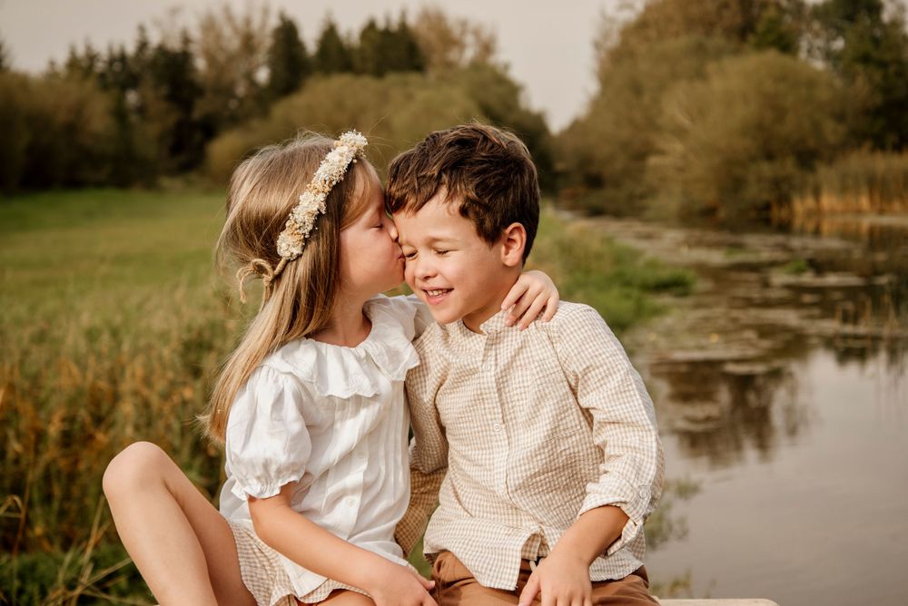 Two children sharing a tender moment by a pond, girl in white dress with floral crown kissing boy's cheek.