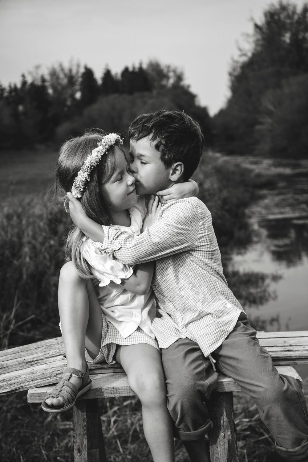 Black and white photo of two young children sharing a sweet moment on a wooden fence near a pond.