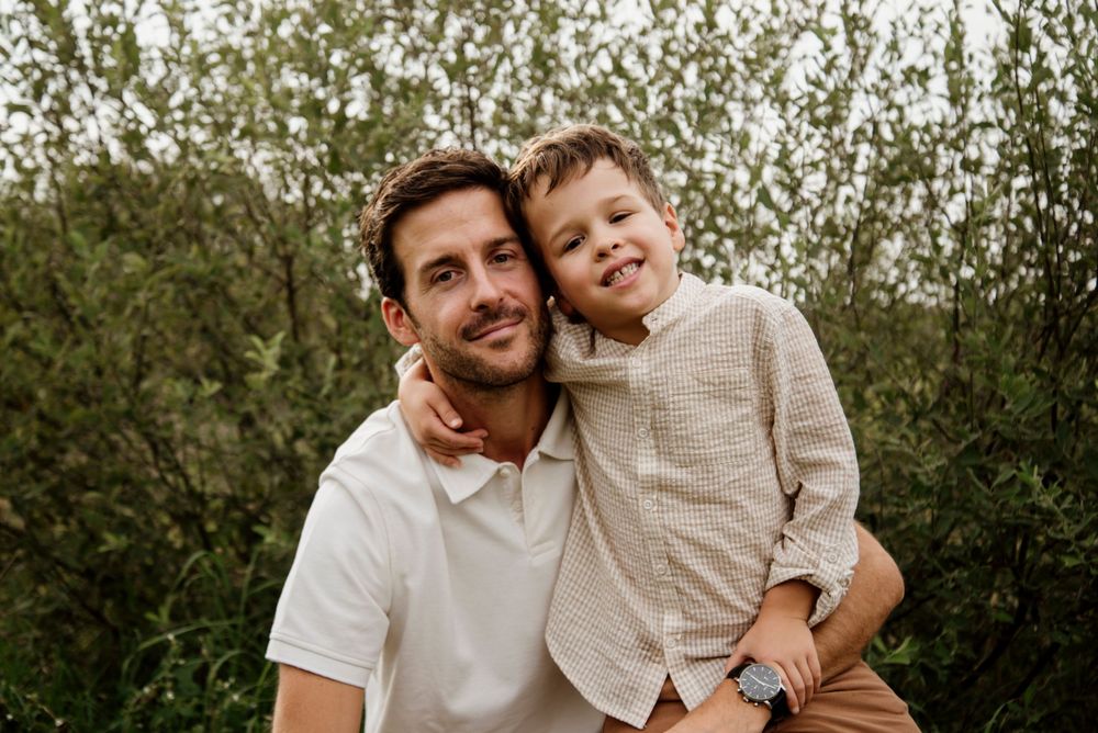 A smiling father holds his young son outdoors, both wearing neutral tones with lush green foliage behind them.