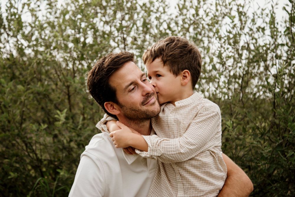 A father holds his young son outdoors, sharing a tender moment as the boy kisses his cheek near lush green bushes.