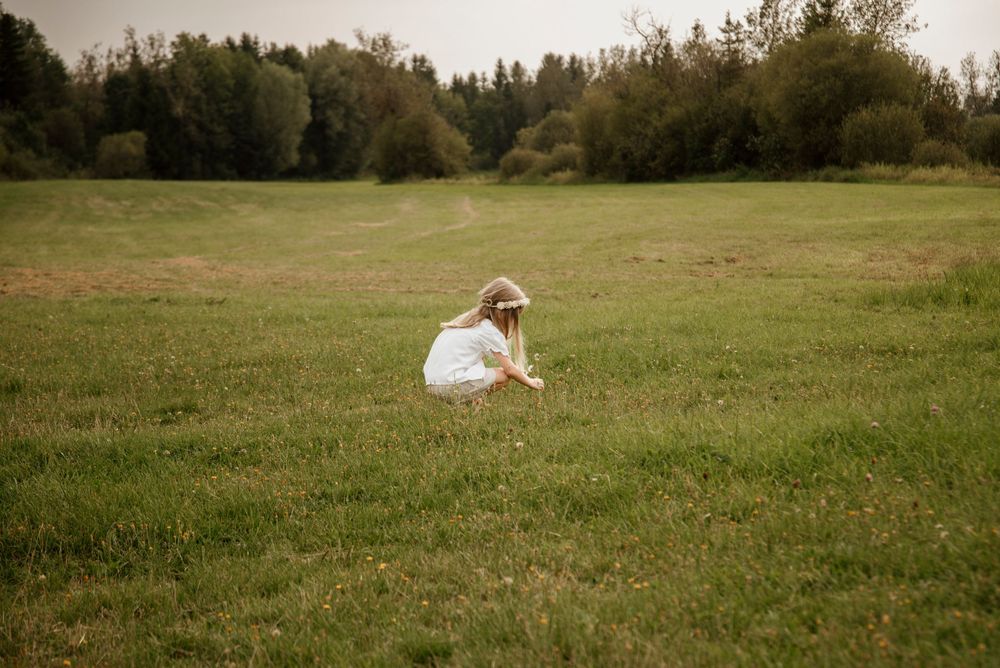 A young girl in a white dress and hat crouches in a green meadow, picking flowers with trees in the background.
