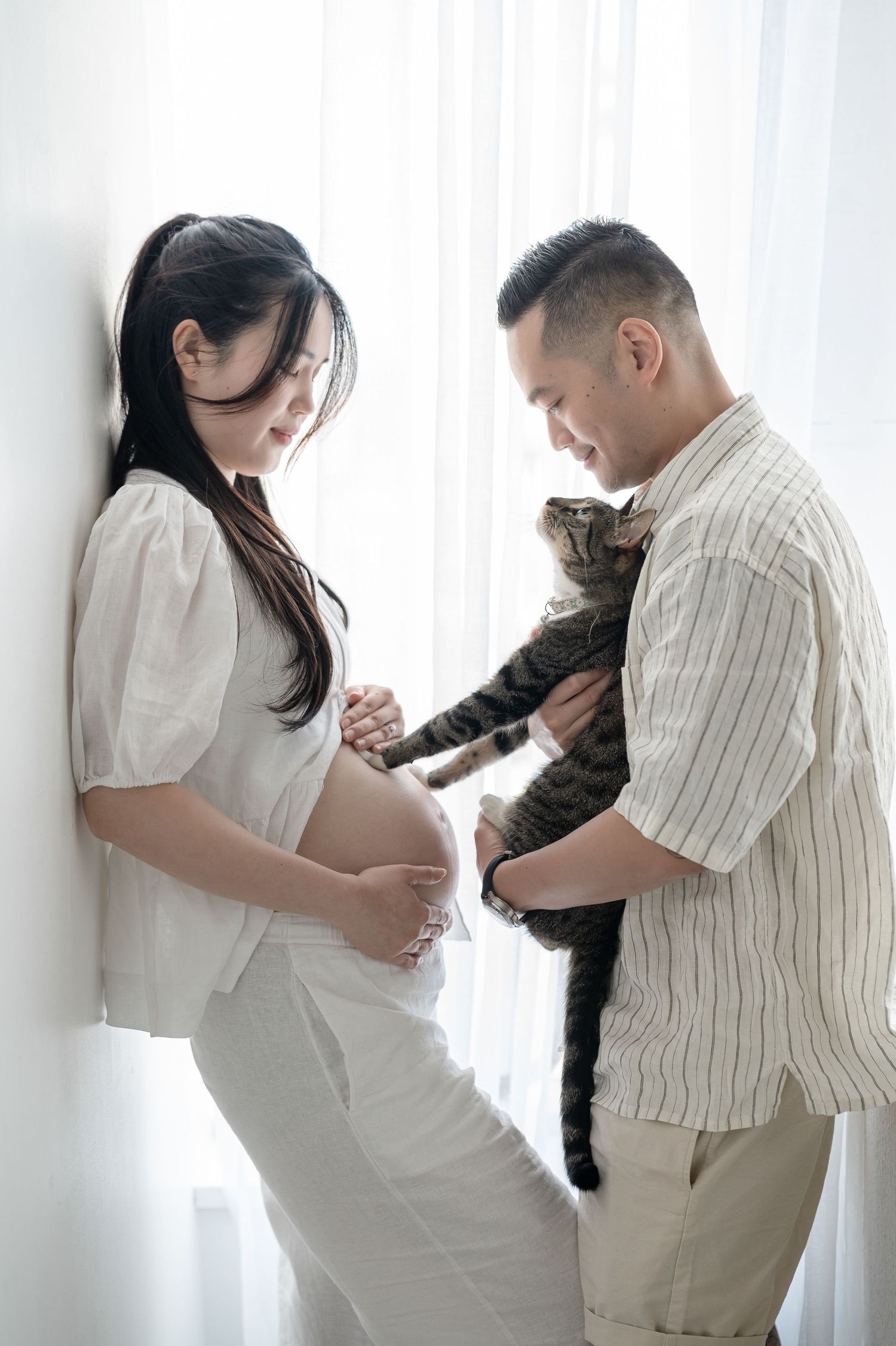 Pregnant Asian woman and man holding a tabby cat together indoors near white curtains.