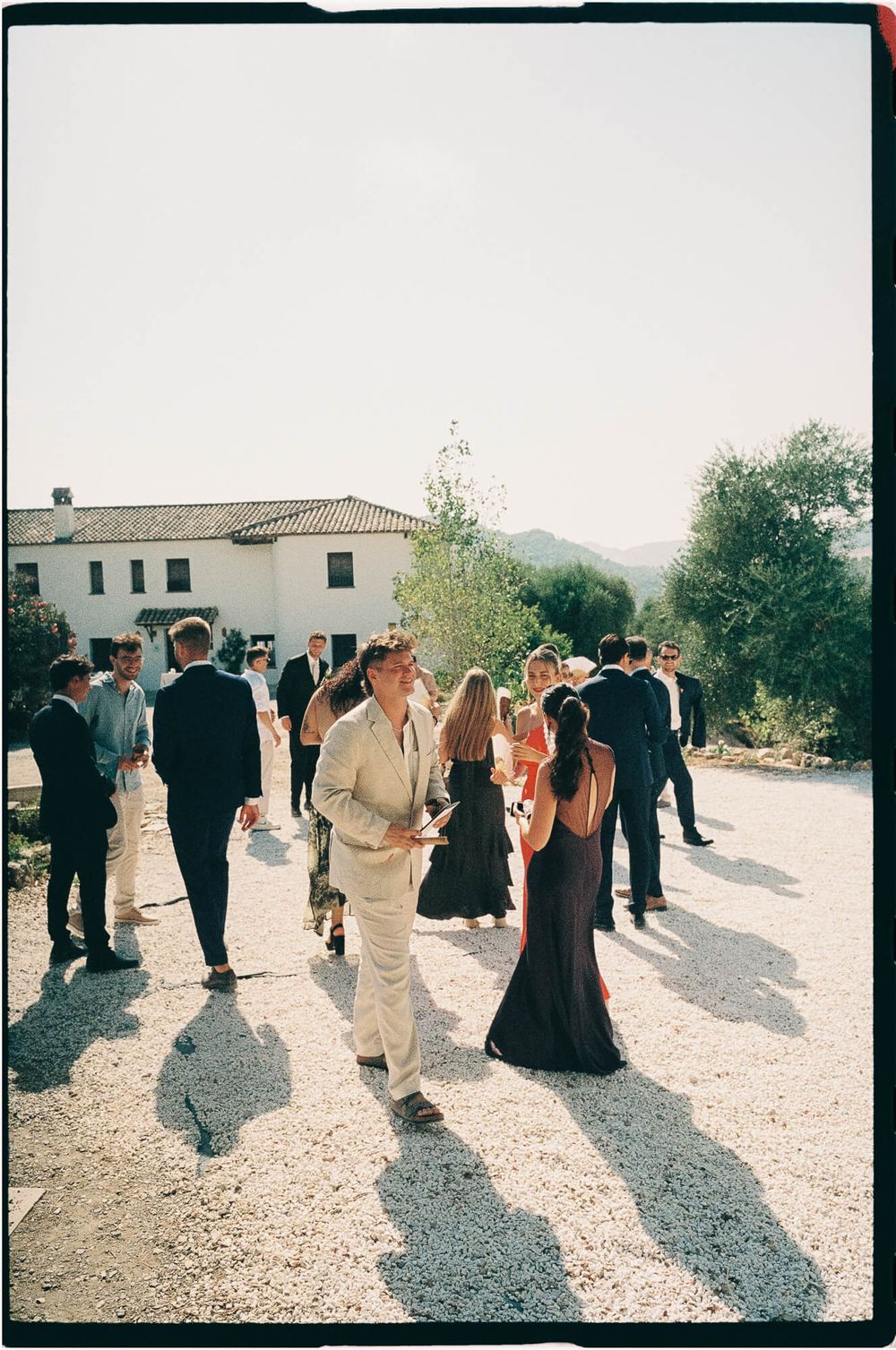 Wedding guests mingle on a sunlit gravel courtyard outside a white Spanish villa with mountain views in the background.