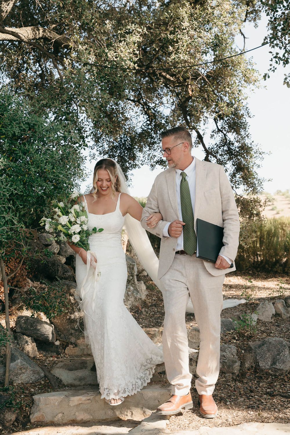 Bride in white lace gown holding bouquet walks with suited man in green tie on stone steps outdoors.