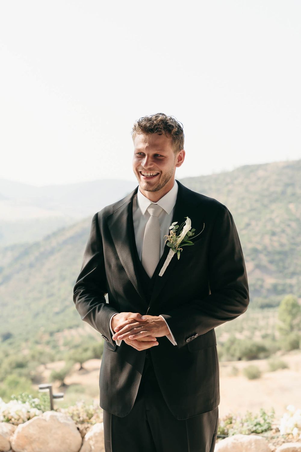 Smiling groom in black tuxedo with floral boutonniere stands before scenic mountain backdrop on wedding day.