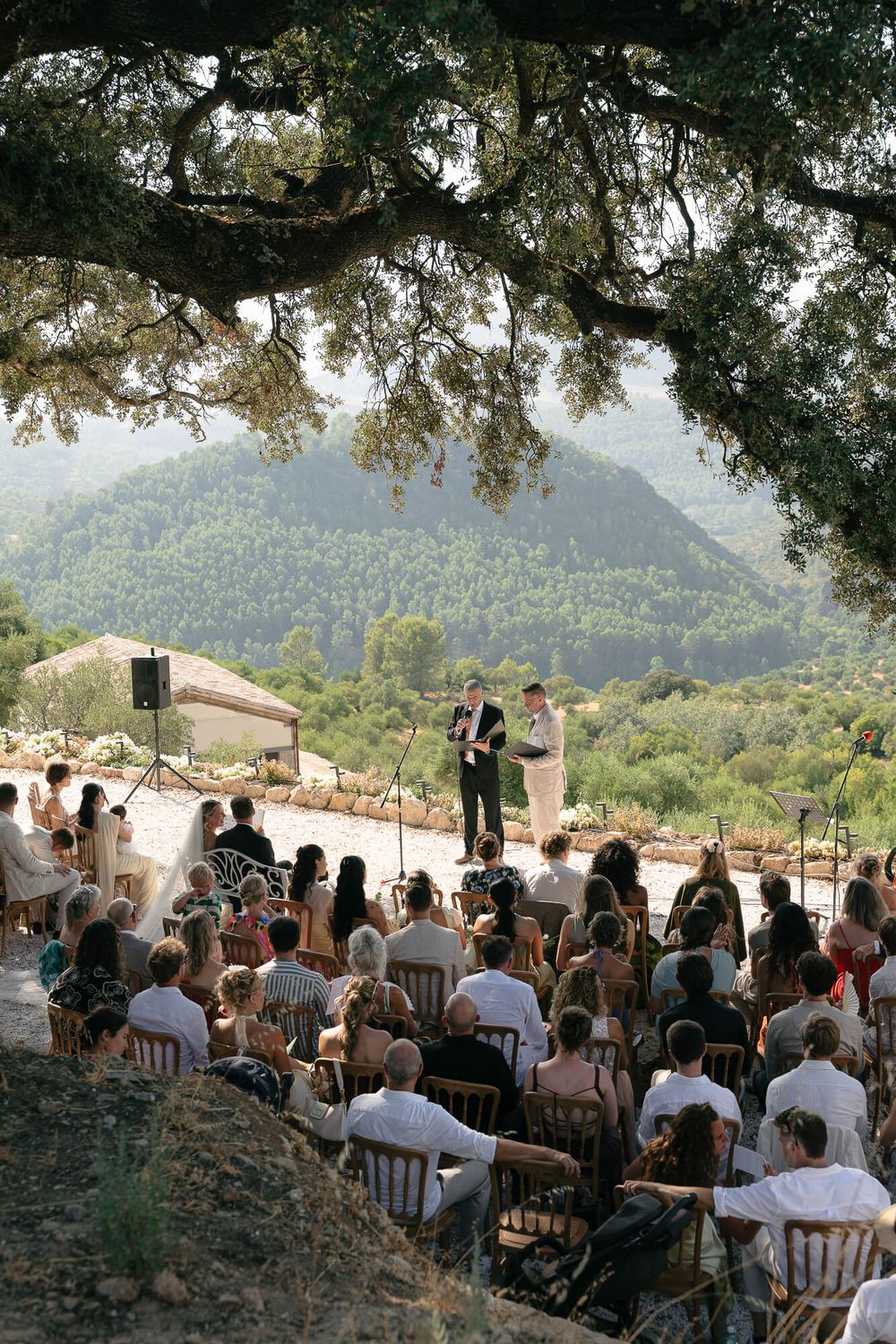 Outdoor wedding ceremony with guests seated under a large tree, overlooking lush green mountains in a scenic Mediterranean landscape.