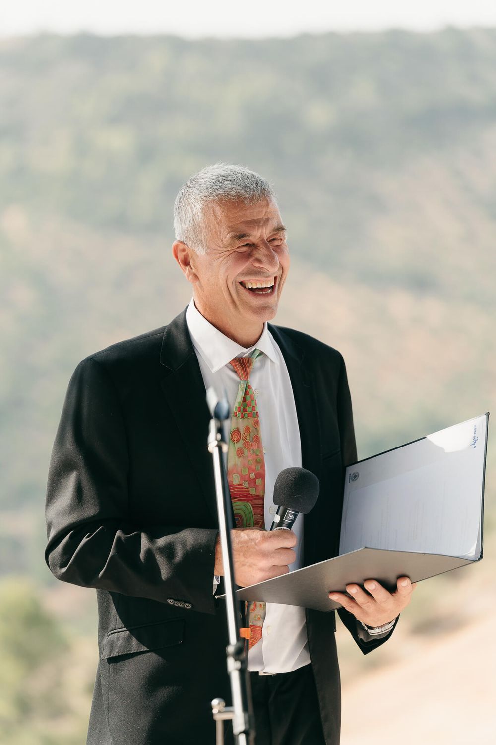 Smiling senior man in black suit with floral tie holding folder and microphone outdoors.