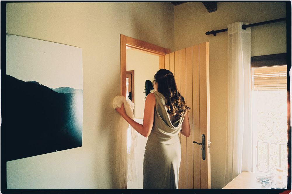 Bride in elegant silver gown holding bouquet, standing before mirror in rustic wedding venue room.