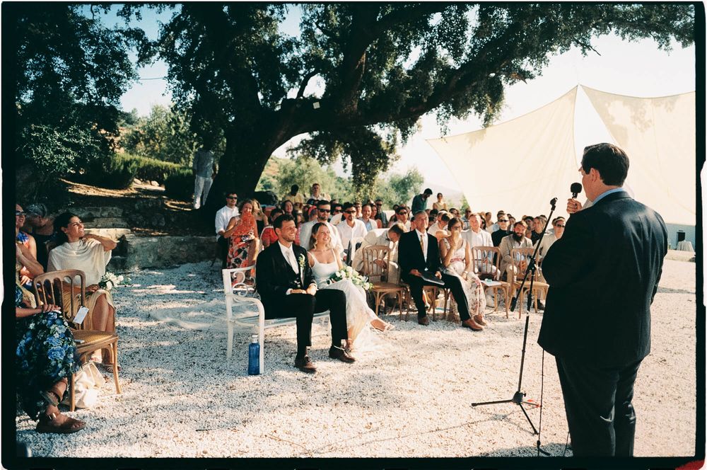 Outdoor wedding ceremony under a large tree with guests seated in rows facing the couple and officiant.