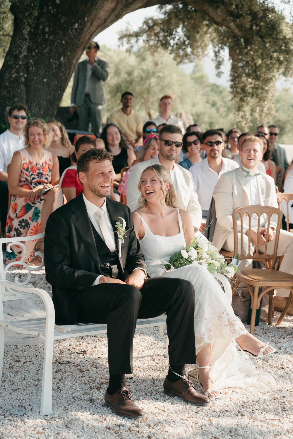 Bride and groom laughing joyfully during outdoor wedding ceremony with guests seated behind them under a large tree.
