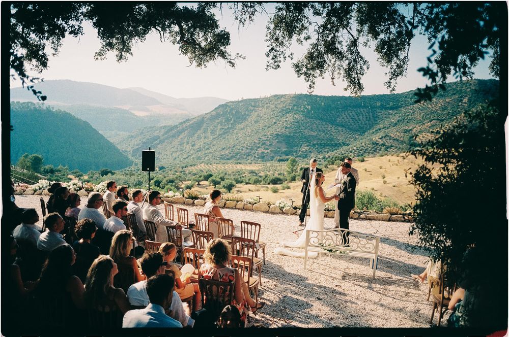 Outdoor wedding ceremony with guests seated overlooking a stunning mountain valley landscape on a sunny day.