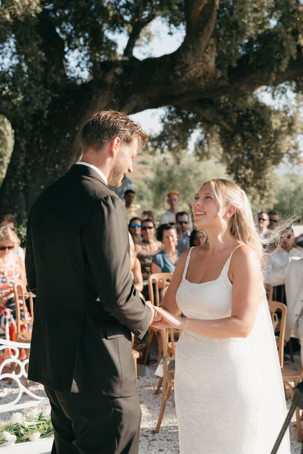 Bride and groom exchange vows outdoors under a large tree during a sunny wedding ceremony with guests seated behind them.