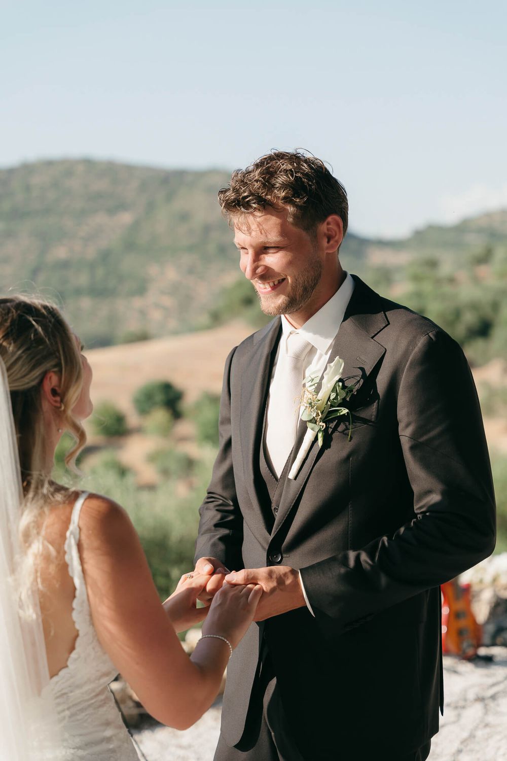 Smiling groom in dark suit placing wedding ring on bride's finger during outdoor ceremony with scenic hills backdrop.