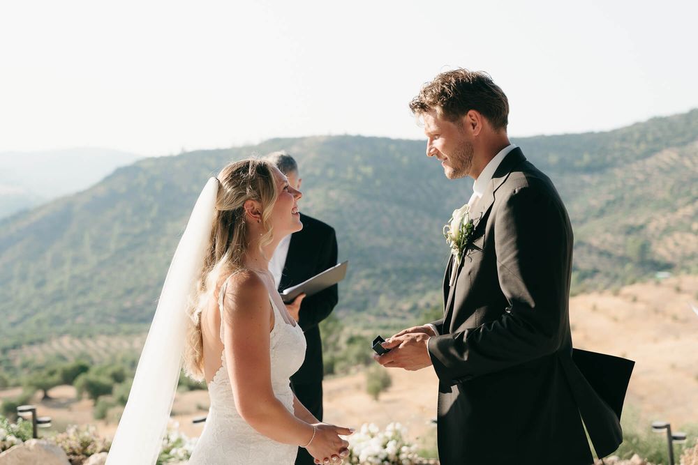 Bride and groom exchange vows outdoors with mountain views, officiant behind them on a sunny wedding day.