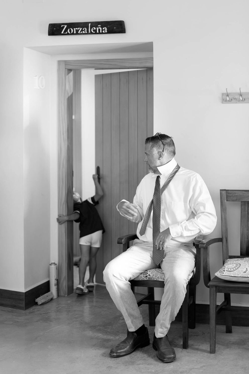Black and white photo of a man in shirt and tie sitting on a chair near a door labeled Zorzalena.