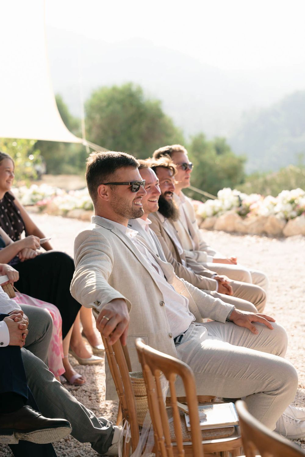 Smiling wedding guests seated outdoors in sunlight, wearing elegant attire, with mountains and greenery in the background.