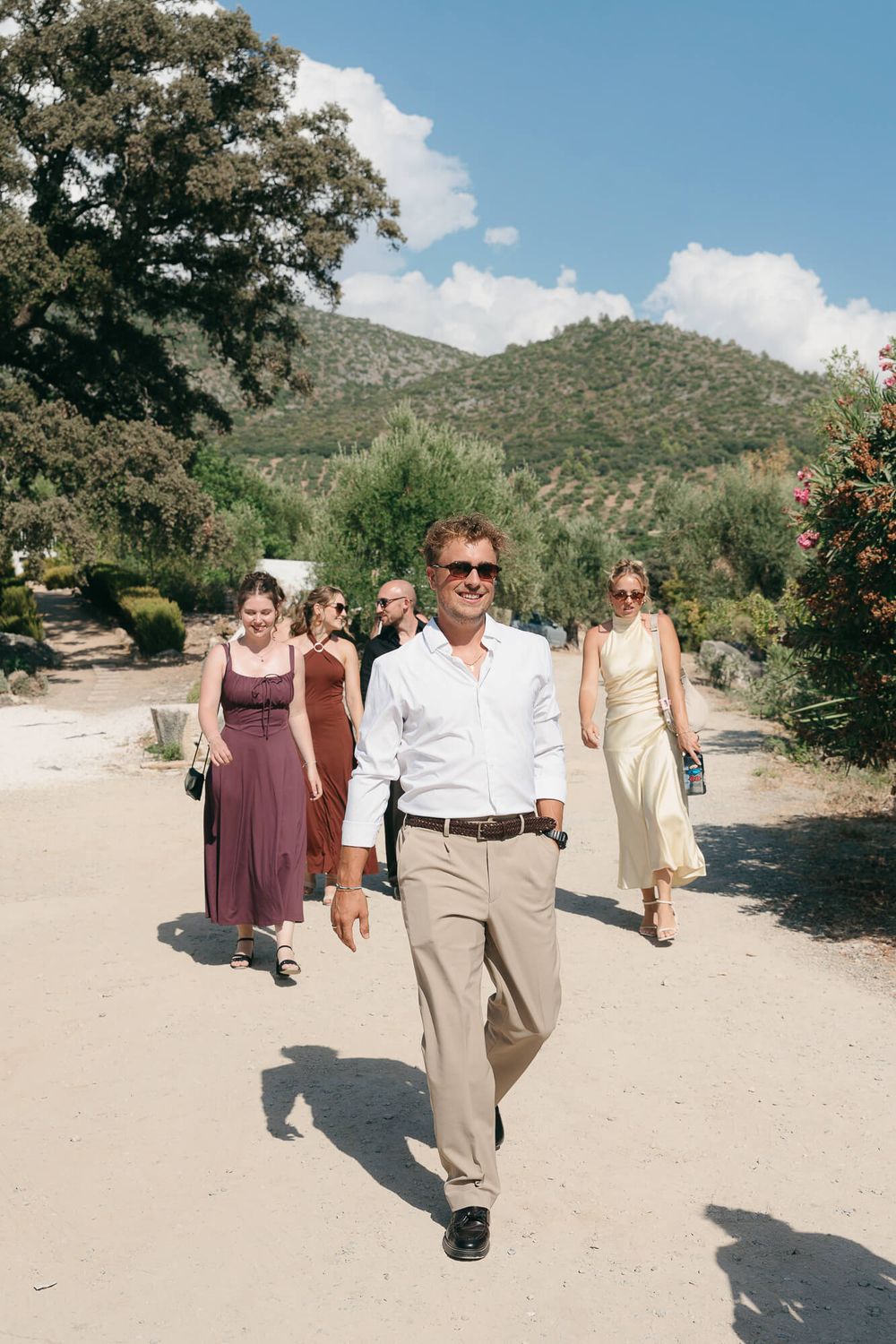 A man in a white shirt and sunglasses leads a group walking along a sunny path with green hills in the background.