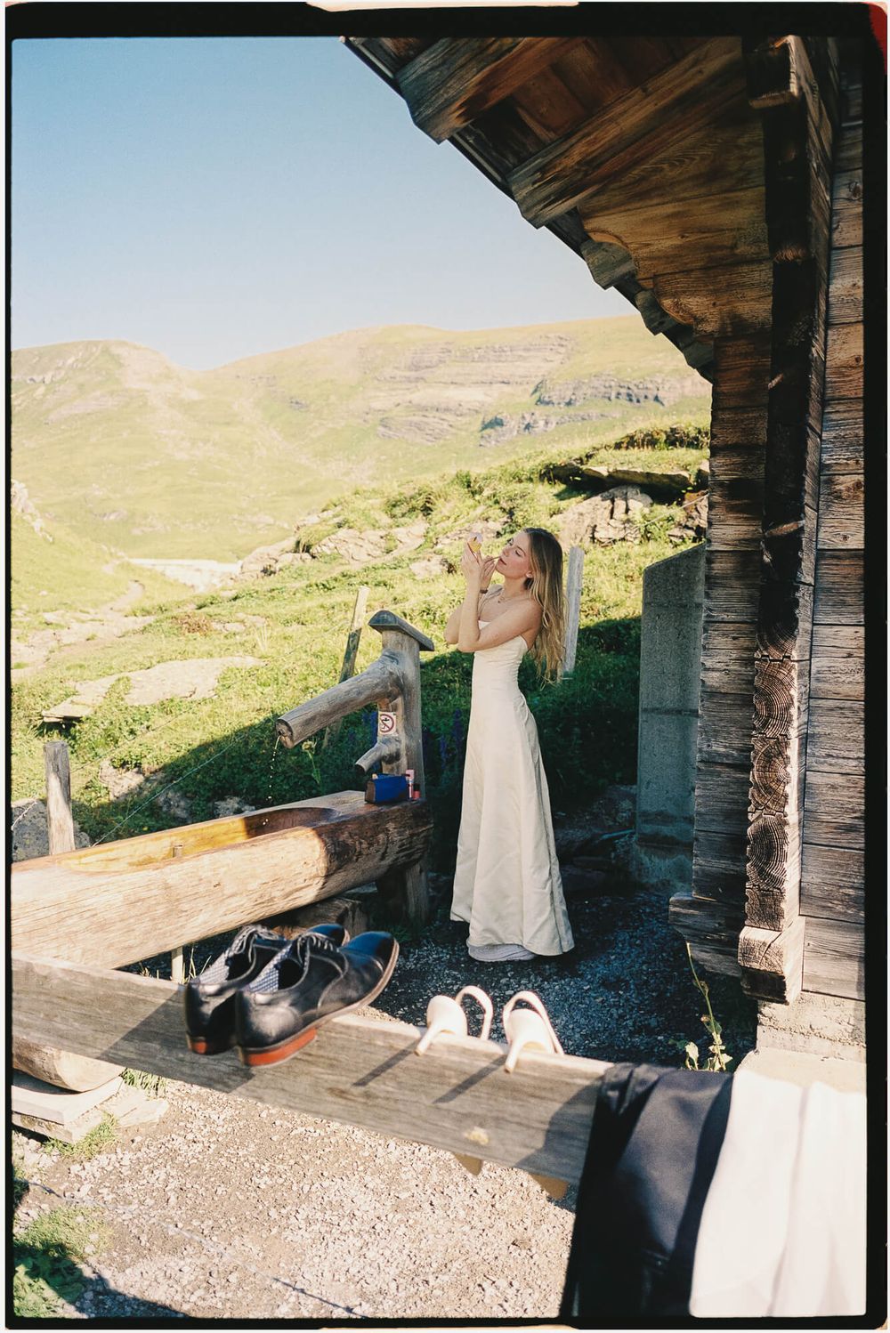 Woman in white dress standing by rustic wooden cabin with scenic green mountain valley backdrop on sunny day.