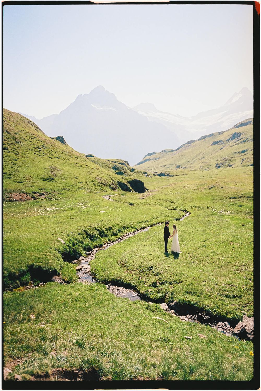 Couple standing by a winding stream in a lush green alpine valley with misty mountain peaks in the background.