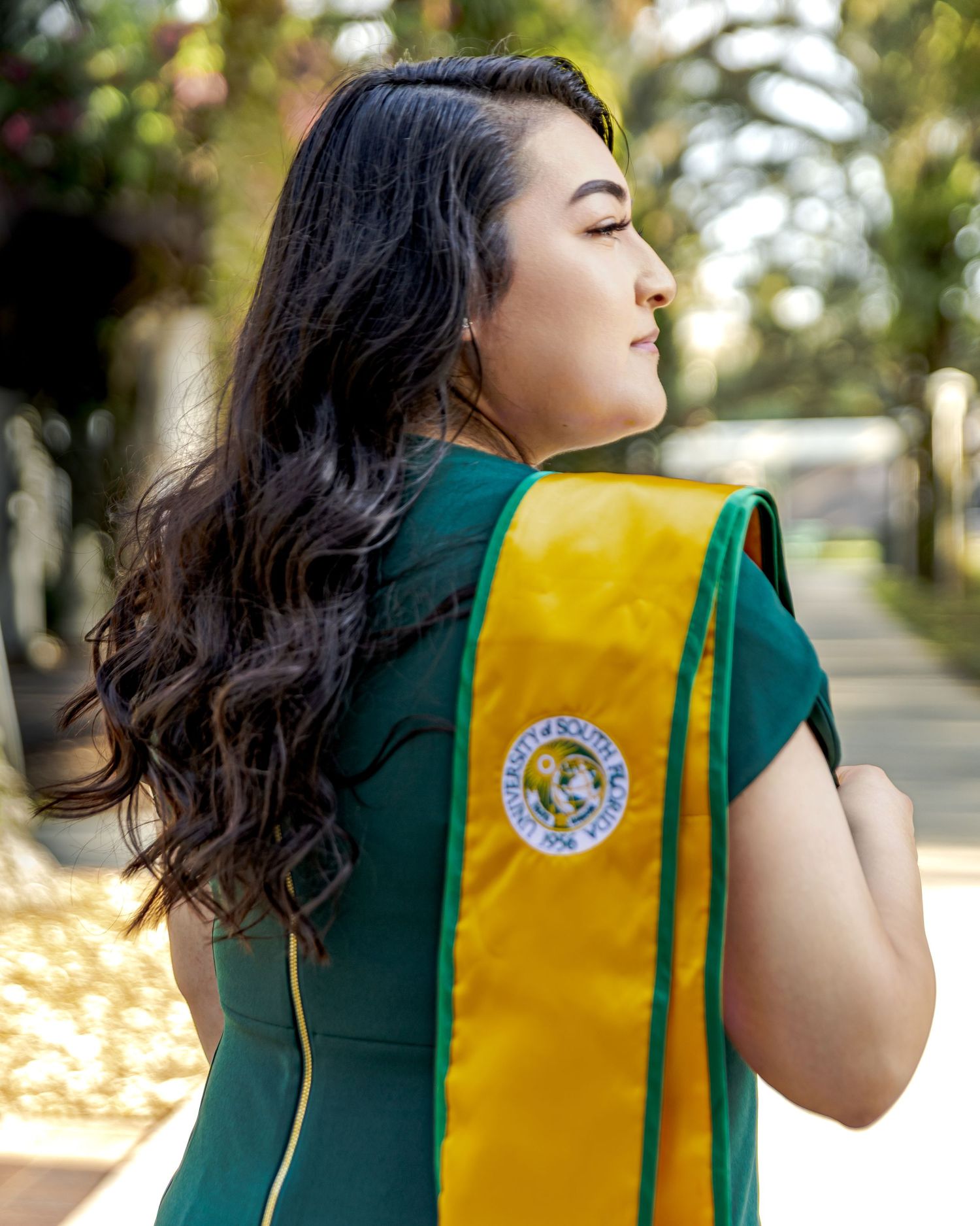 Young woman in green dress wearing gold graduation stole with university seal, posed outdoors on campus.