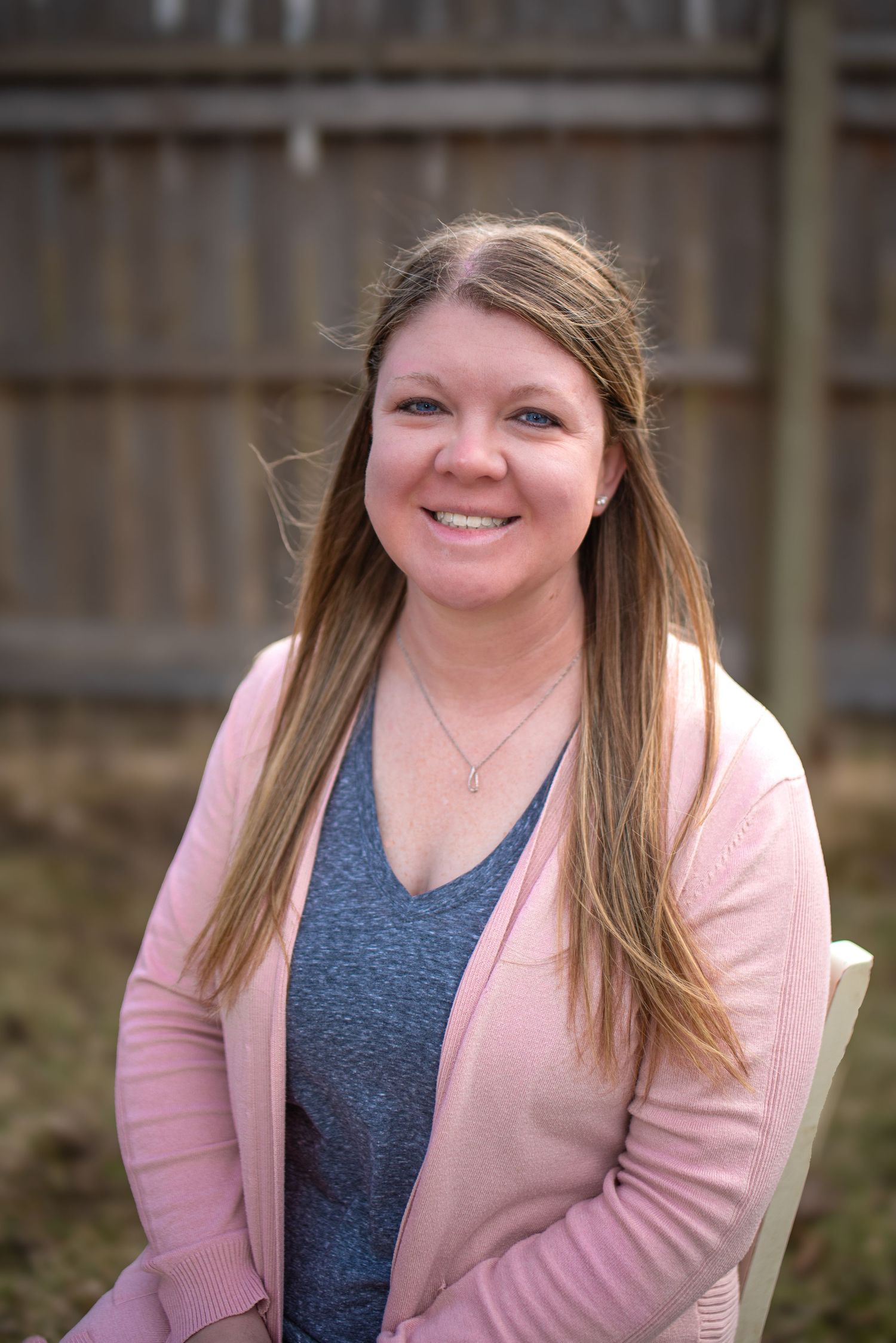 Smiling woman with long brown hair wearing a pink cardigan and gray top, seated outdoors near a wooden fence.
