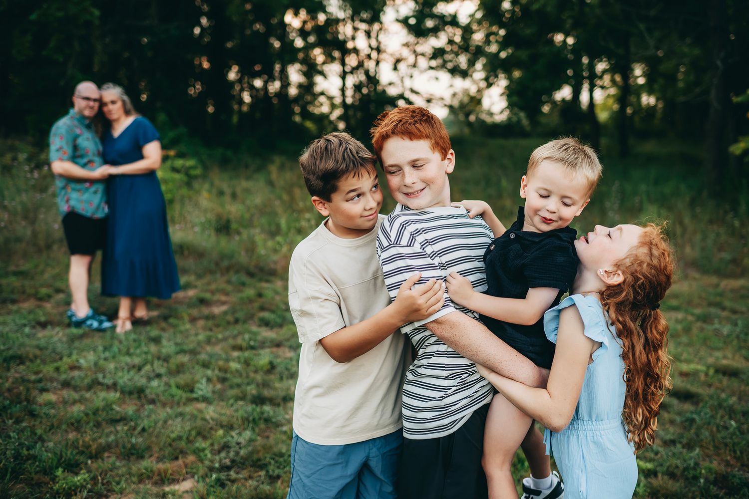 Four children playfully embrace outdoors while a couple shares a tender moment in the background at a family photo session.