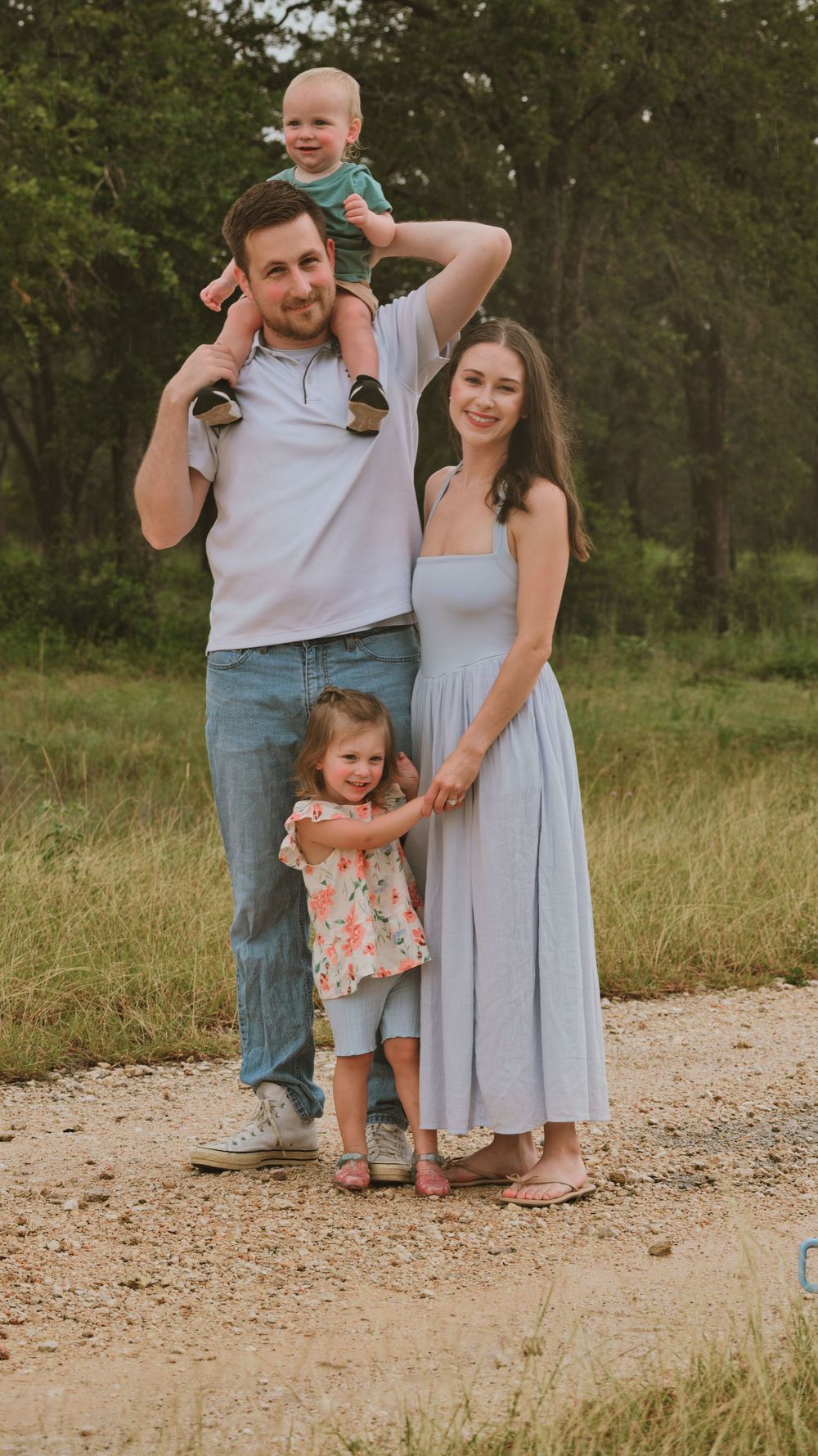 Young family of four poses on a gravel path, dad holding toddler on shoulders with mom and daughter beside him.