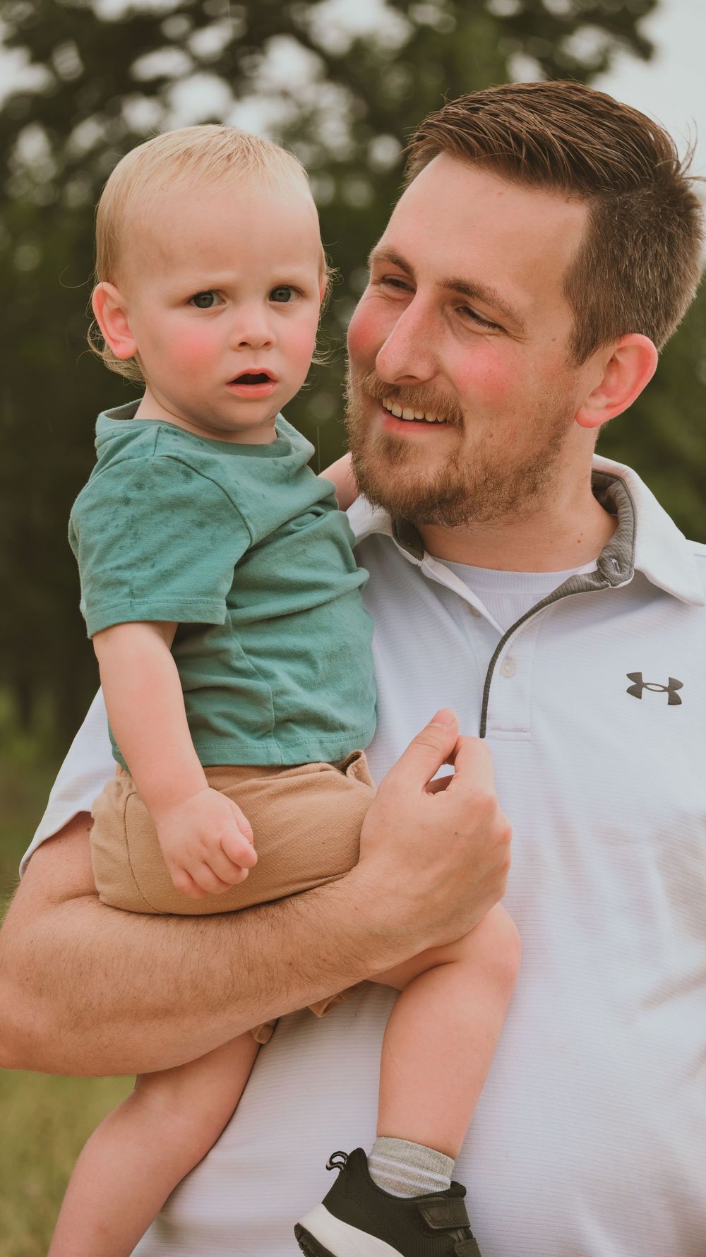 A smiling bearded man in a white Under Armour shirt holds a blonde toddler boy in a green shirt outdoors.