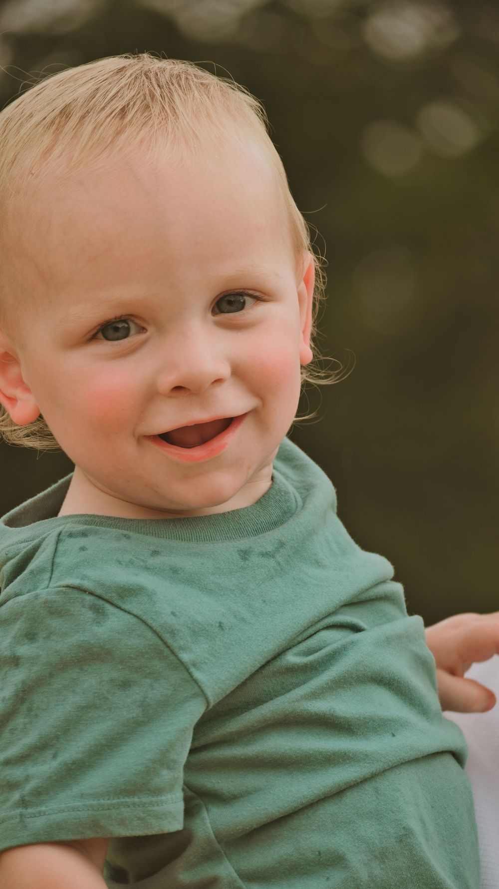 A smiling toddler with blonde hair wearing a green t-shirt poses outdoors with a blurred green background.