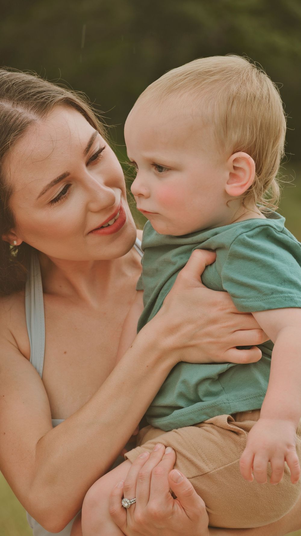A smiling mother holds her blonde toddler boy in a green shirt outdoors.
