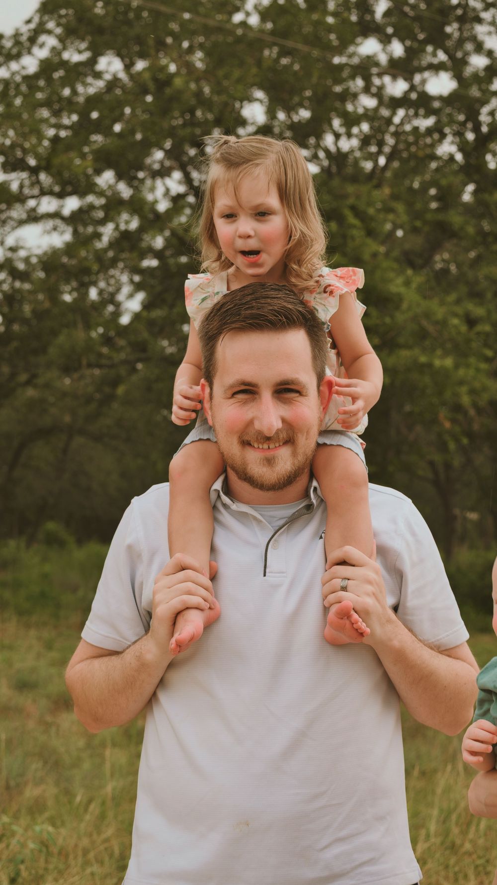 A smiling man carries a young girl on his shoulders outdoors, surrounded by lush green trees.
