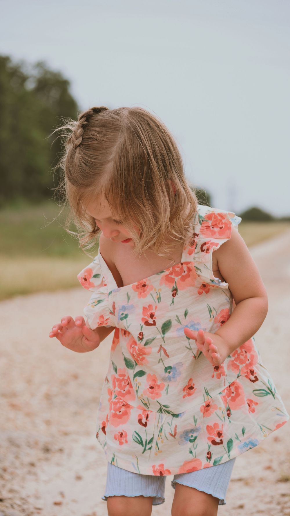 A toddler girl in a floral dress plays on a sandy path outdoors.