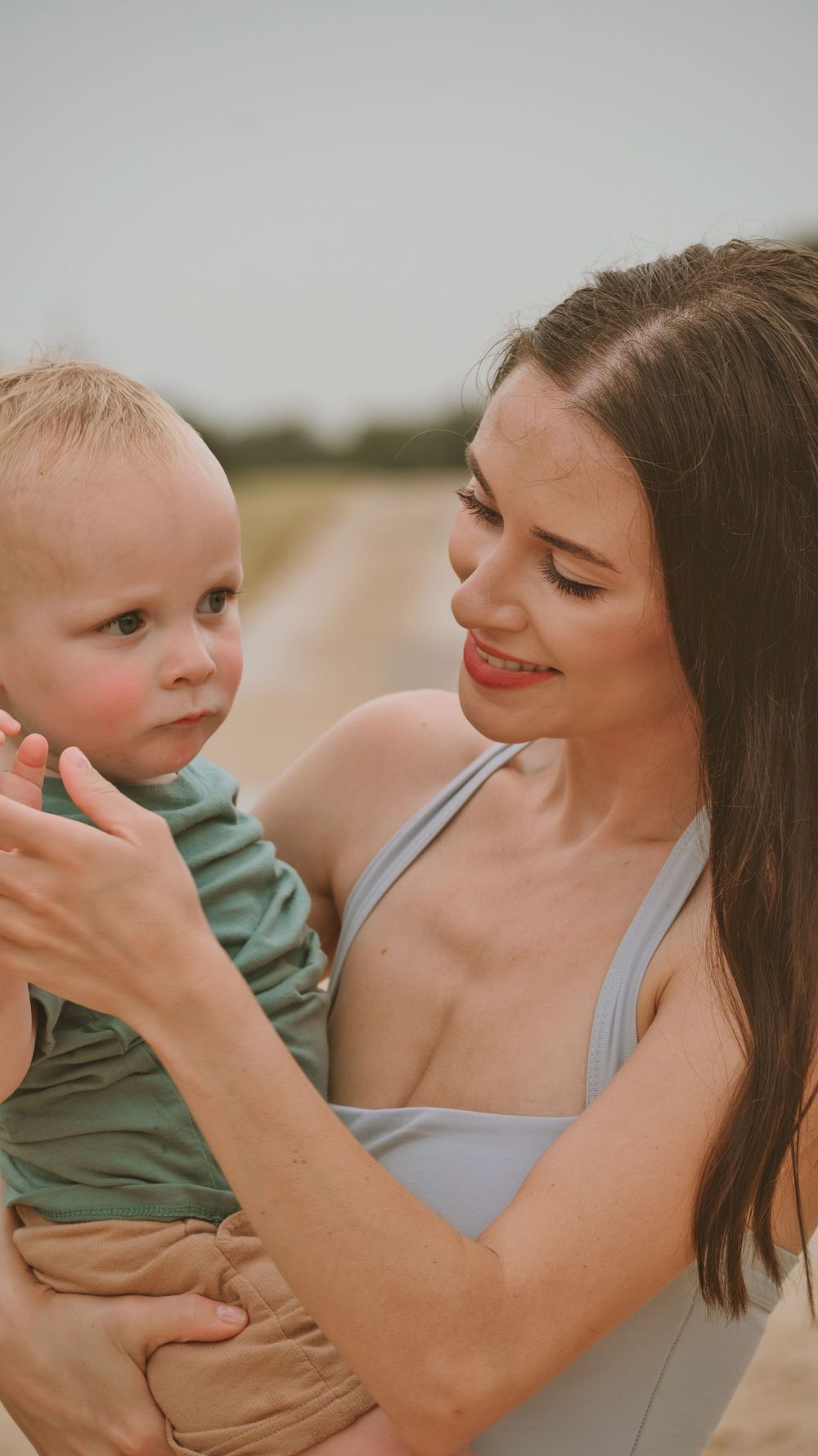 Loving mother in gray dress holding and smiling at her blonde toddler boy outdoors.