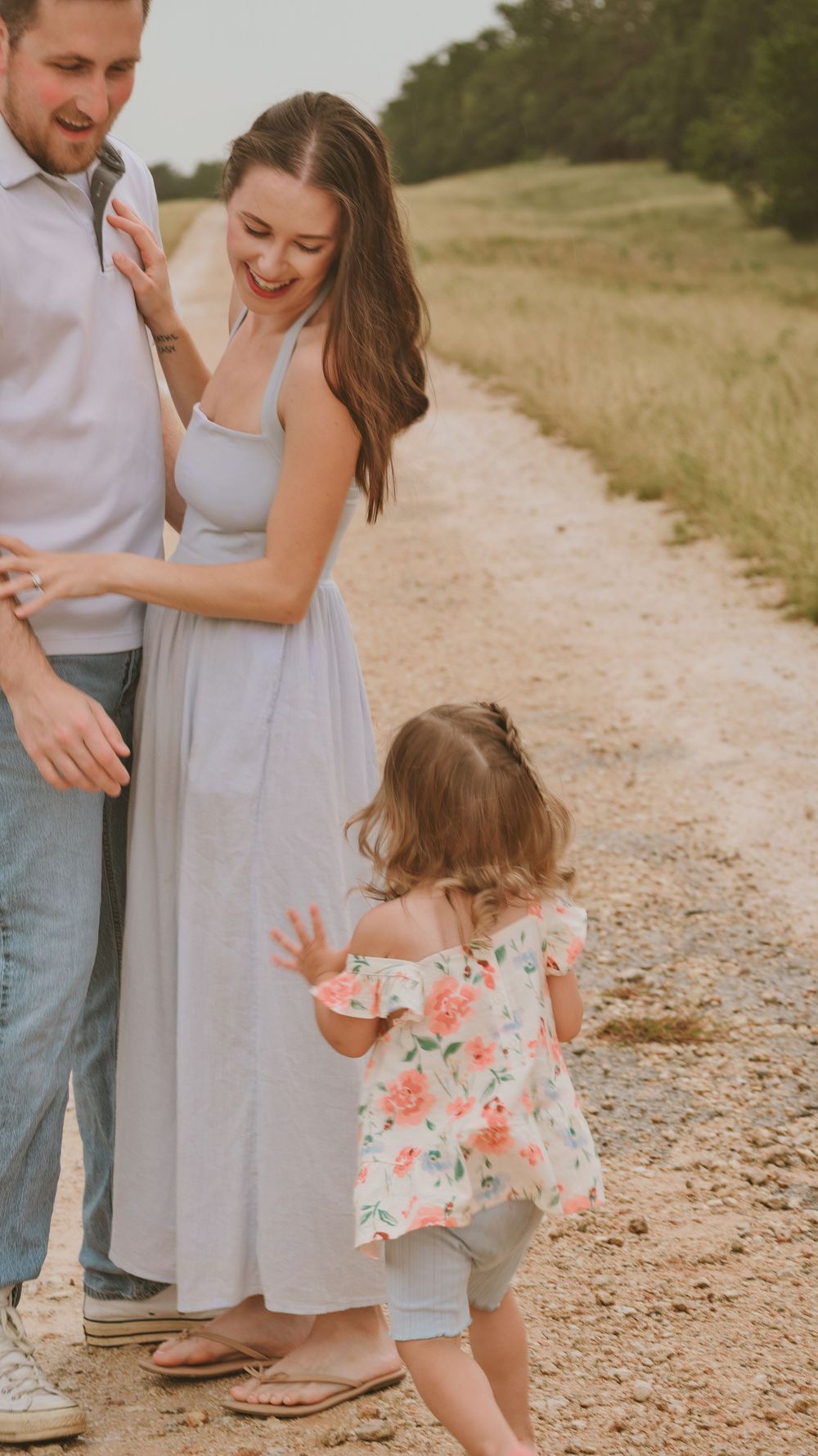 Happy couple smiles together on a sunny gravel path as their young daughter in a floral dress reaches up to them.
