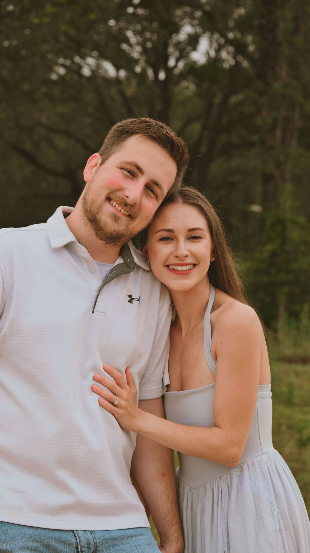 Smiling couple posing outdoors in a wooded setting, man in white polo and woman in gray dress embracing.