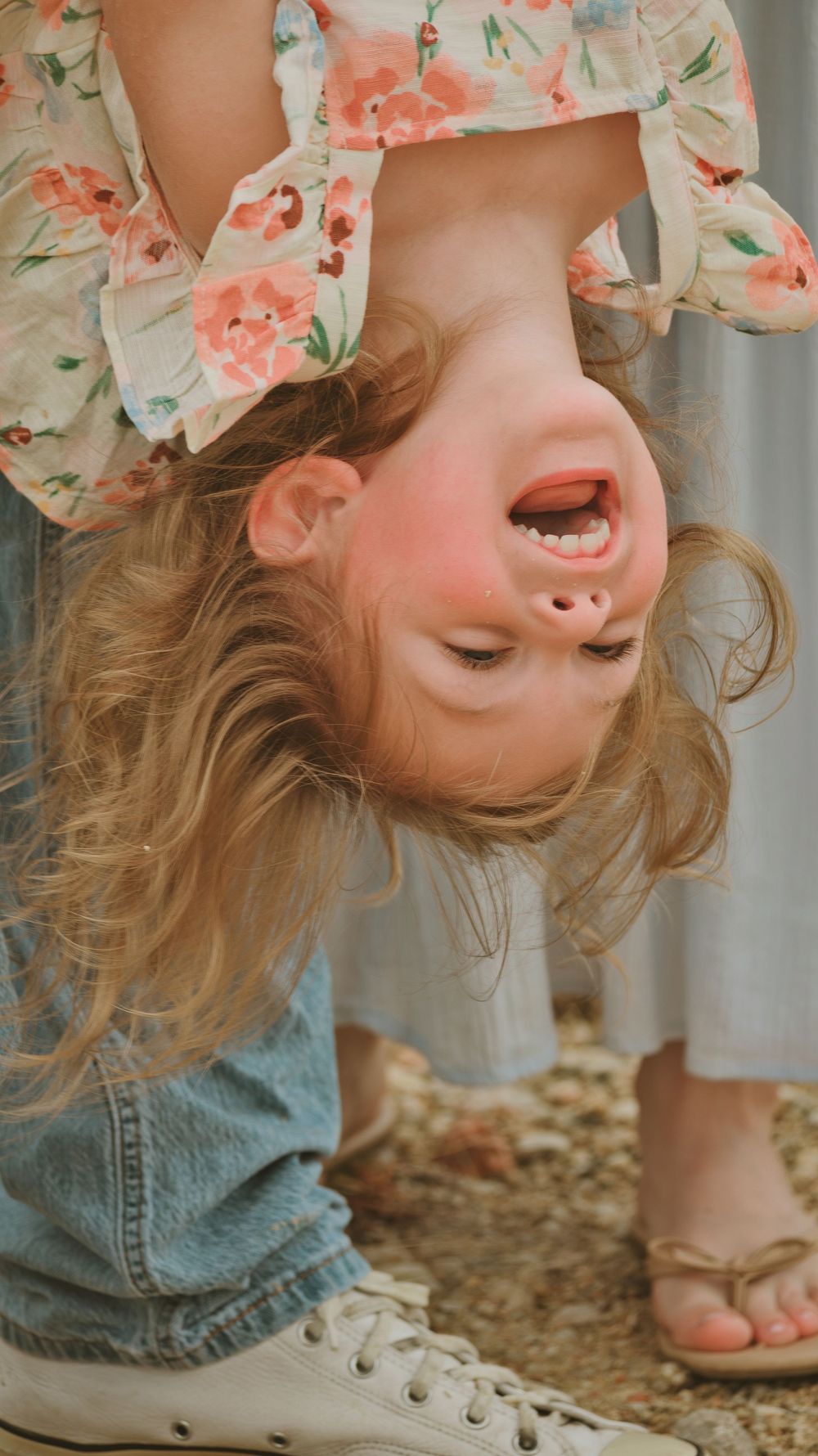 A laughing toddler girl held upside down, wearing a floral top and jeans, with rosy cheeks and blonde hair.