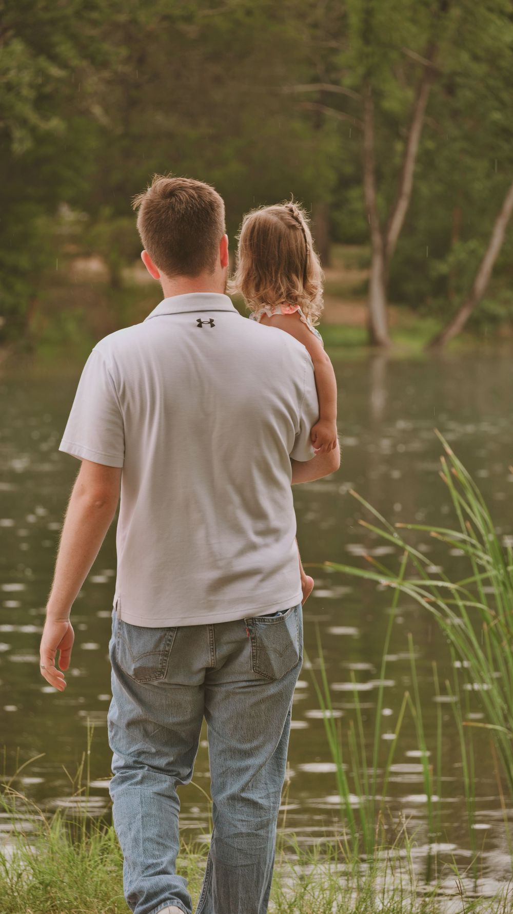 Father carrying young daughter while walking near a serene pond outdoors.