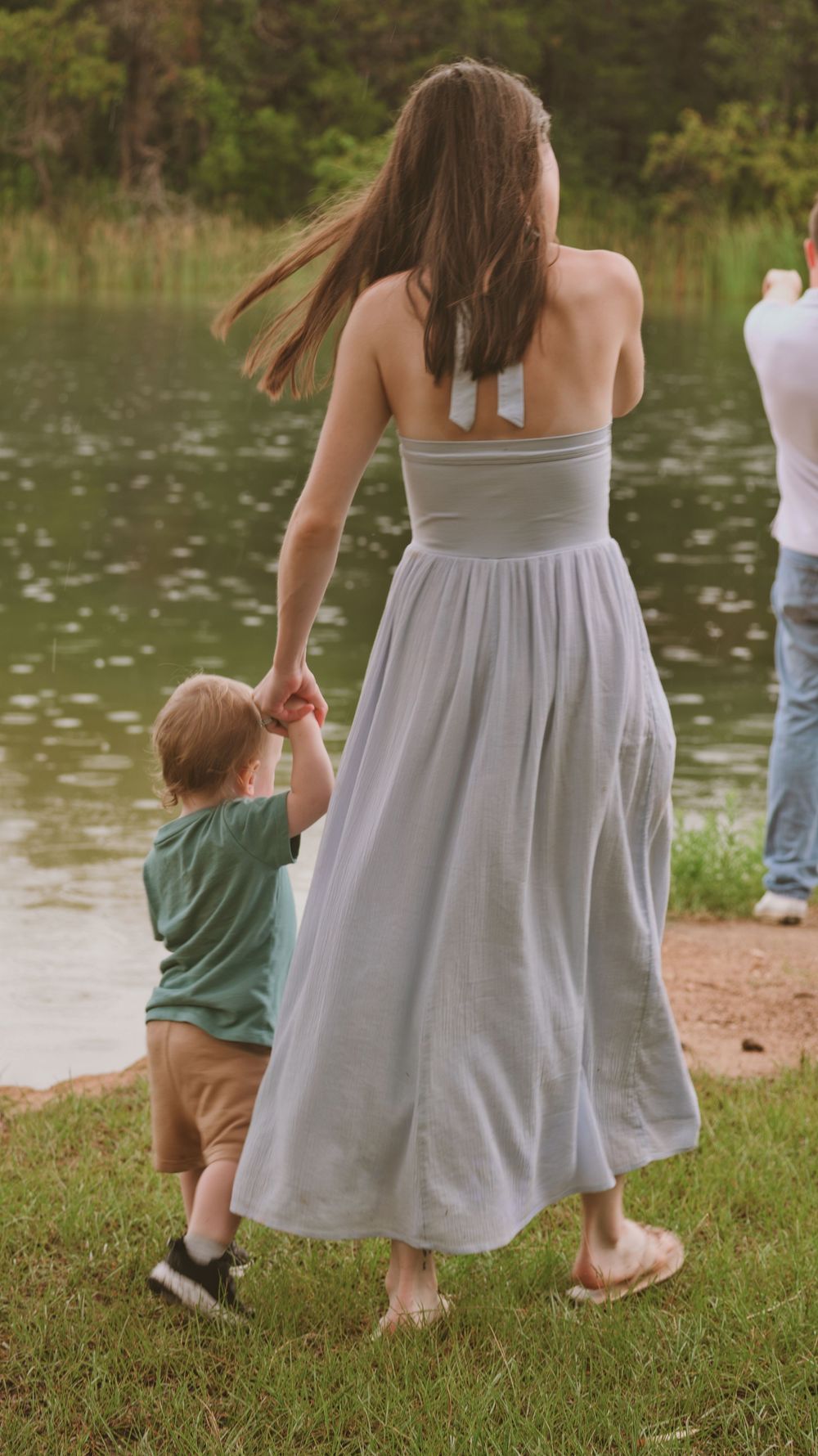Mother in gray maxi dress holding young son's hand by a peaceful lake.