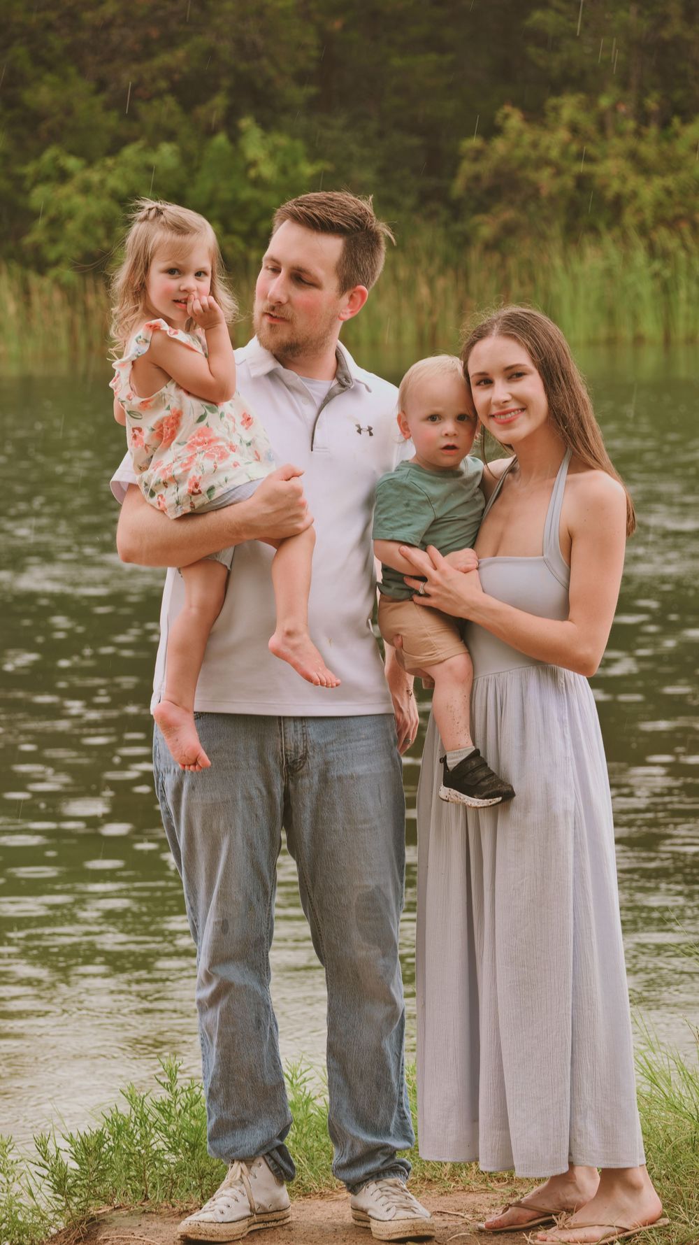 A young family poses by a lake, parents each holding a small child.