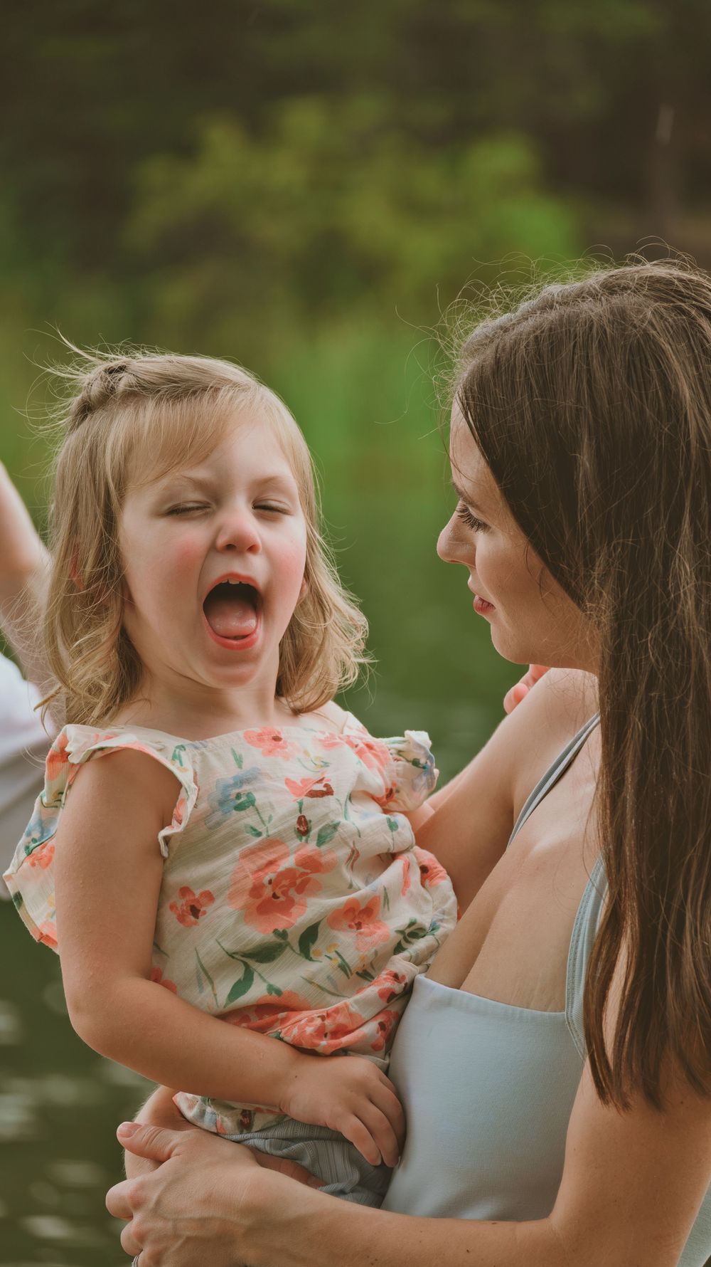 A mother holds her expressive toddler daughter in a floral dress outdoors near a lake.