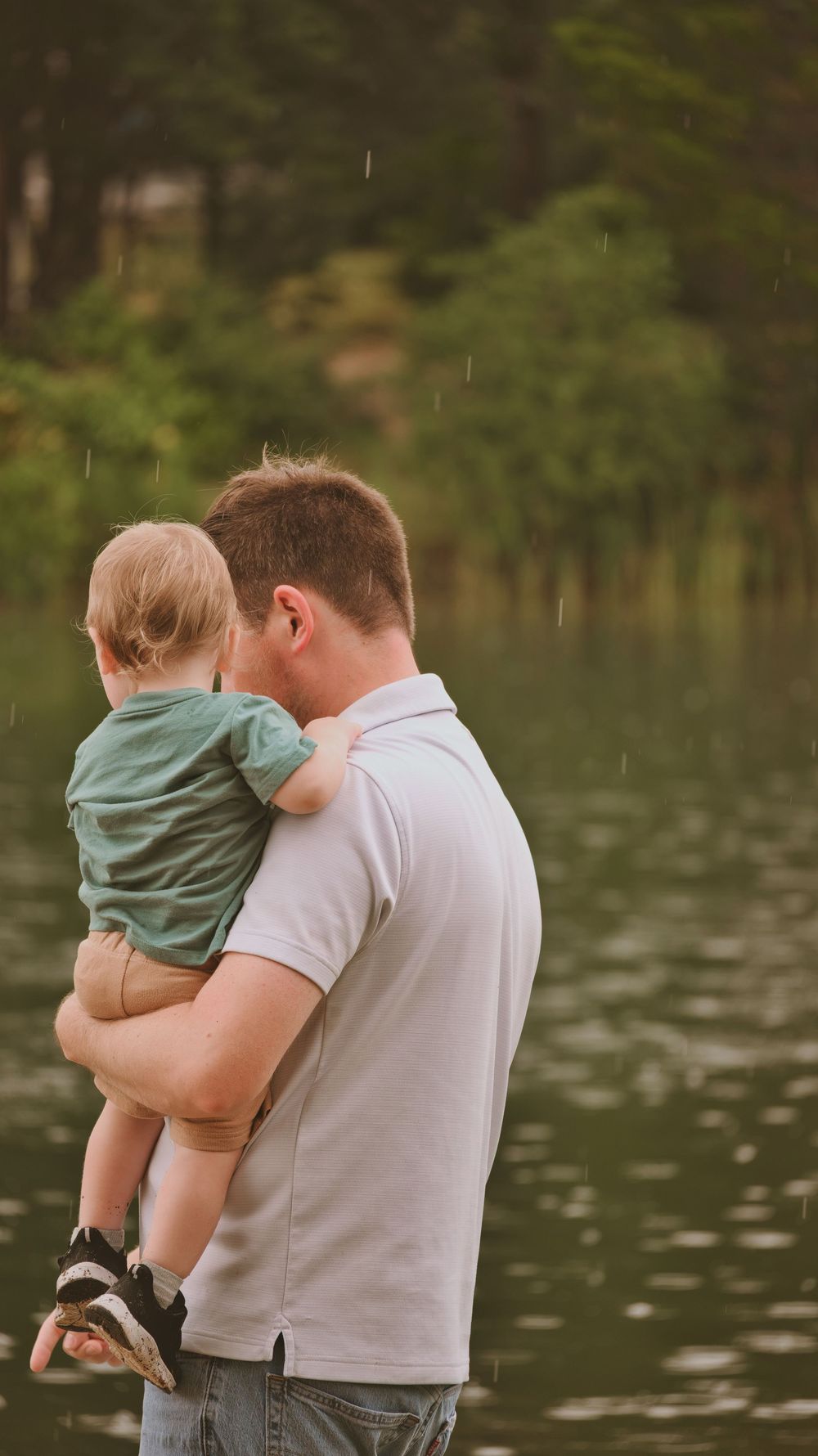 A father tenderly holds his young baby son near a serene lake in the rain.