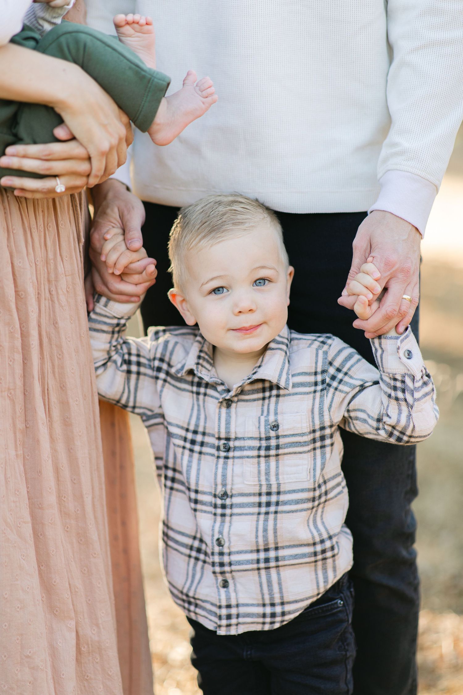 A blonde toddler boy in a plaid shirt holds his parents hands during an outdoor family photo session.