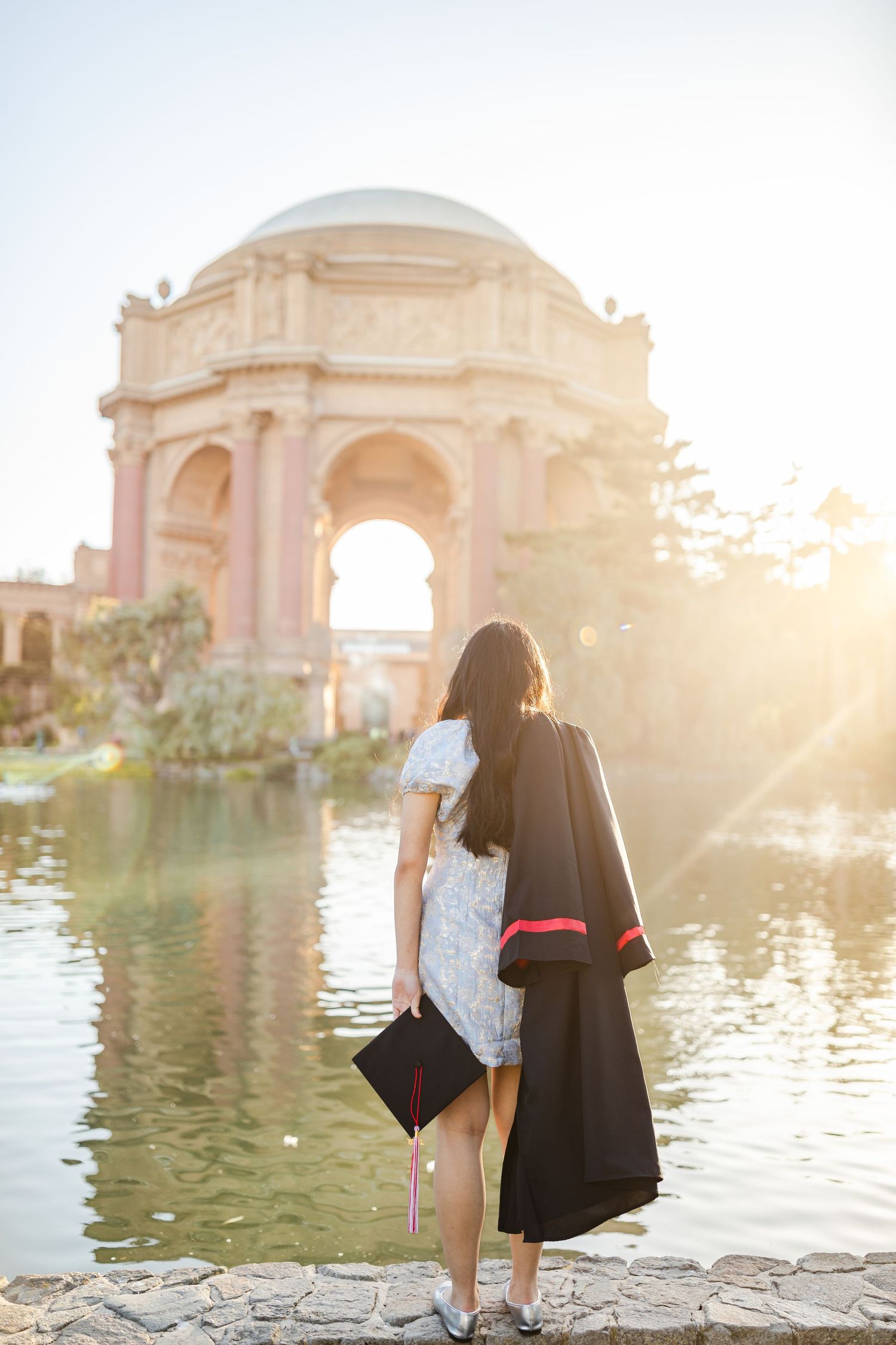 Graduate holding cap and gown by a sunlit lagoon with the Palace of Fine Arts rotunda in San Francisco.